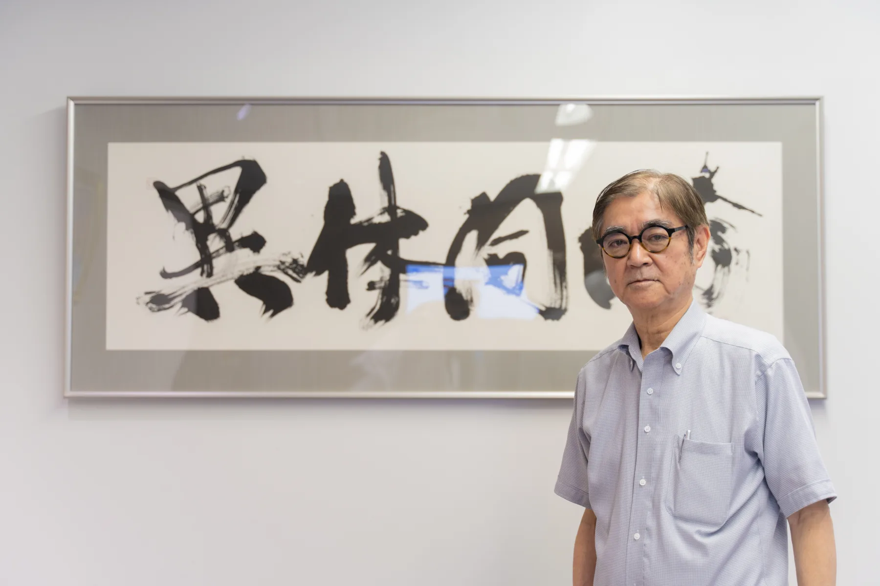 書作品を背に立ち正面を見据える小城雅弘